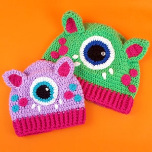 May include: Two crocheted hats with large eyes and pink ears. The hats are green and purple with pink trim.