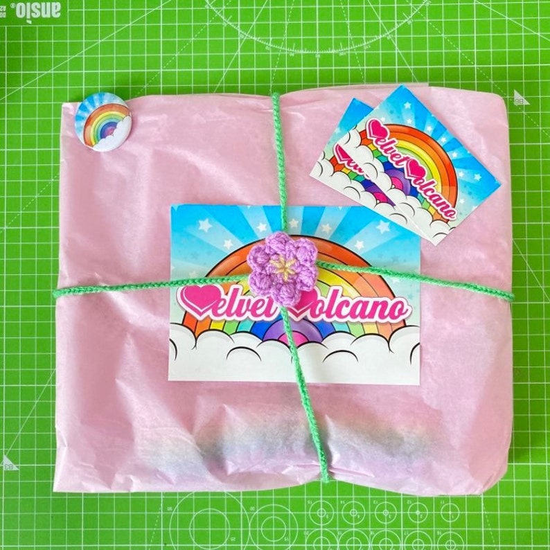 Pu&ograve; includere: Un pacchetto rosa avvolto in carta velina con uno spago verde. Il pacchetto ha un adesivo con un arcobaleno e il testo "Velvet Volcano". C'&egrave; un piccolo fiore all'uncinetto sul pacchetto.