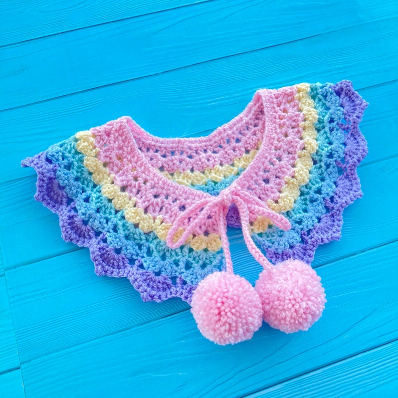 May include: A rainbow crochet collar with a tie closure and two pink pom-poms attached. The collar is on a blue wooden background.