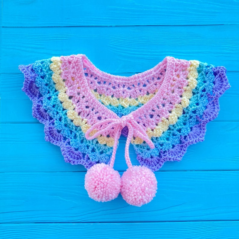 May include: A rainbow crochet collar with pink pom-poms attached to the ends of the ties. The collar is on a blue wooden background.