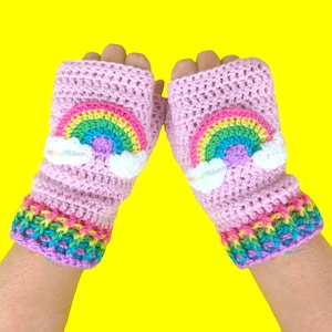 May include: A pair of pink fingerless gloves with a rainbow and cloud design. The gloves are crocheted and have a colorful border.