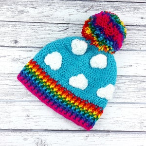 May include: A colourful crochet beanie hat. The hat has a bright blue crown with white cloud appliqués, a rainbow-striped brim, and a large, multi-coloured pom-pom. The hat is made of soft yarn, perfect for keeping warm in the colder months.