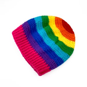 May include: A rainbow crochet beanie hat with a pink brim. The hat is made with different coloured yarn, including red, orange, yellow, green, blue, indigo, and violet.