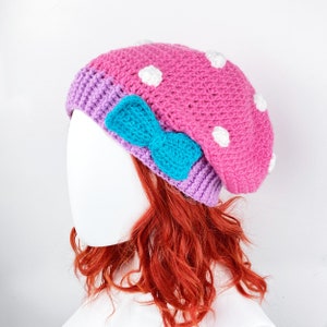 Può includere: Un basco rosa all'uncinetto con pois bianchi e un fiocco turchese. Il cappello ha un bordo viola ed è esposto su una testa di manichino bianca. È un accessorio fatto a mano.