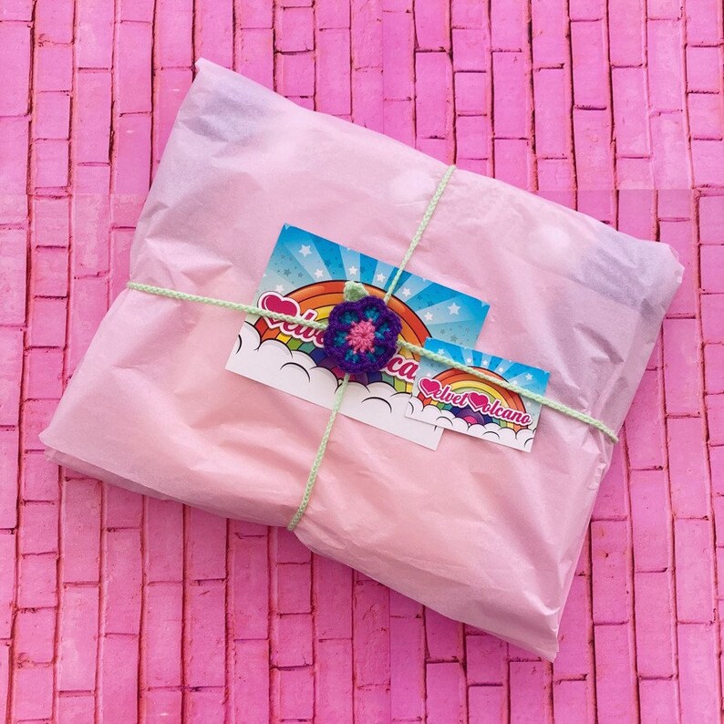 May include: A pink and white gift wrapped in tissue paper with a small card that says "Velvet Volcano" and has a rainbow and a purple flower.