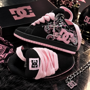 May include: Black and pink DC sneakers featuring thick pink laces, a pink DC logo on the side, and a silver cross charm attached to the eyelets. The sneakers have a black suede upper with pink graphic patterns and a matching black and pink box.