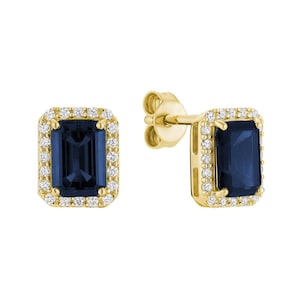 May include: A pair of yellow gold stud earrings featuring emerald-cut dark blue centre stones. Each rectangular gemstone is surrounded by a halo of small, round, clear white diamonds set in a yellow gold frame with post back closures.