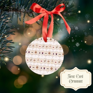 Puede incluir: Un adorno navideño redondo de color blanco con un patrón repetitivo de varias ilustraciones de cartas del tarot, que incluyen soles, lunas, leones y espadas. El adorno está rematado con un lazo de satén rojo brillante para colgar.