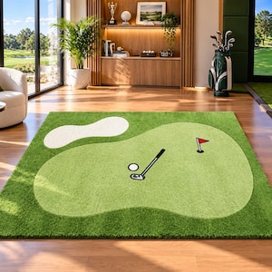 Op de afbeelding: Een rechthoekig vloerkleed met een golfbaanontwerp, voorzien van een lichtgroene putting green, een witte zandbunker, een golfclub, een witte golfbal en een hole met een rode vlag. Het kleed heeft een textuur die op gras lijkt.