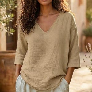May include: A beige V-neck top with three-quarter length sleeves and a relaxed fit. The garment features a textured fabric surface and a straight hemline, worn tucked into light-coloured trousers. The design is simple with no patterns.