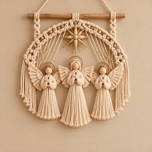 Könnte beinhalten: Ein Wandbehang mit drei Makramee-Engeln mit Holzköpfen und goldenen Heiligenscheinen, die an einem Holzstab hängen. Das Design umfasst einen zentralen Stern, komplexe Knotenmuster und Holzperlen, alles gefertigt aus cremefarbenem Baumwollseil.