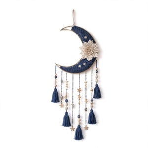 Pode incluir: Um pendente de parede em forma de meia-lua com fio de crochê azul-escuro enrolado em uma moldura. É decorado com uma grande flor de tecido na cor creme, pequenos detalhes de estrelas brancas e vários fios pendentes de contas, pingentes de estrela e borlas azuis.