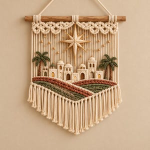 May include: A macrame wall hanging featuring a cream-colored cord design of a desert town with domed buildings, two palm trees, and a central star. The piece is suspended from a wooden dowel and includes layered green and brown rope accents.