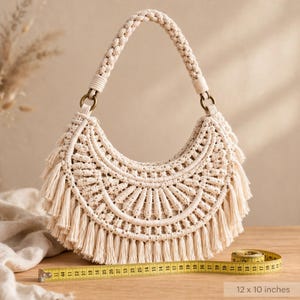Pode incluir: Uma bolsa de macramé em forma de meia-lua na cor branco sujo, com padrões de nós intrincados, uma alça trançada fixada por anéis em tom de bronze e franjas na borda inferior. Uma fita métrica amarela mostra medidas de 30,48 x 25,4 cm.