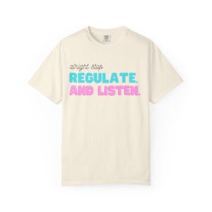 Op de afbeelding: Een crèmekleurig T-shirt met korte mouwen en de tekst "alright stop" in zwart cursief schrift, gevolgd door "REGULATE." in dikke cyaan blokletters en "AND LISTEN." in dikke roze blokletters, gecentreerd op de voorzijde van het kledingstuk.