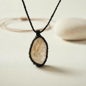 May include: A necklace featuring a raw, translucent, pale yellow crystal pendant encased in a black macrame cord setting. The pendant hangs from a thin, braided black cord, showcasing the natural, irregular shape and textured surface of the stone.