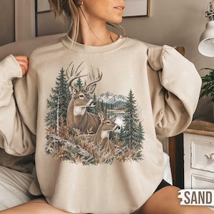 May include: A sand-coloured crewneck sweatshirt featuring a graphic print of a large buck with antlers and a smaller deer standing in a forest landscape with pine trees, autumn foliage, a calm lake, and distant mountains under a clear sky.