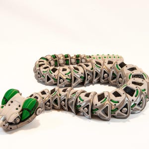Puede incluir: Una figura de serpiente mecánica con un cuerpo segmentado con detalles en blanco, plata y verde brillante. El diseño articulado consta de placas triangulares repetitivas y patrones geométricos intrincados, creando una apariencia robótica.