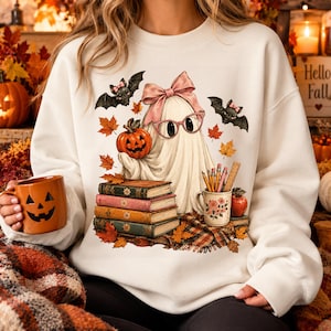 May include: A white crewneck sweatshirt featuring a graphic of a ghost wearing pink glasses and a pink bow, holding an orange pumpkin. The design includes a stack of four books, two bats, autumn leaves, a mug with pencils, and a red apple.