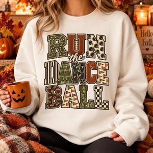 Peut inclure: Un sweat-shirt à col rond couleur crème arborant le texte RUN the DANCE BALL en grandes lettres majuscules à motifs variés. Les lettres intègrent divers designs sur le thème du football, incluant des schémas de jeu et des textures.