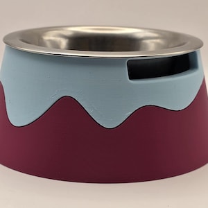 May include: A pet bowl featuring a stainless steel interior and a two-tone exterior base. The base has a maroon bottom section and a light blue top section with a wavy, drip-like pattern. There is a rectangular cutout near the top rim.