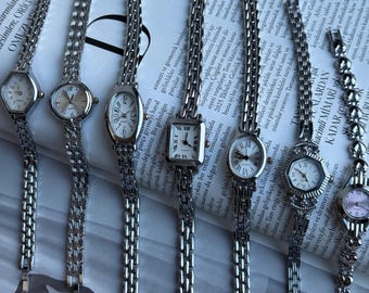 May include: A collection of seven silver-toned wristwatches with metallic link bands. The watches feature various case shapes including round, rectangular, and hexagonal designs with white or light-coloured analogue dials and black hour markers.