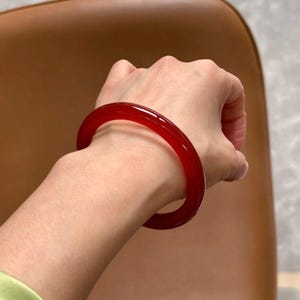 Peut inclure: Un bracelet jonc circulaire et lisse, fabriqué dans un matériau rouge profond translucide. Le bracelet présente une finition polie et brillante qui reflète la lumière sur sa surface arrondie, créant un aspect cramoisi vibrant et uniforme.