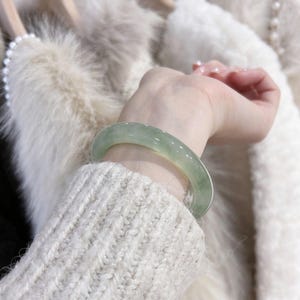 Könnte beinhalten: Ein glatter, durchscheinender hellgrüner Jade-Armreif mit polierter Oberfläche. Das kreisförmige Band weist subtile Farb- und Klarheitsvariationen auf und präsentiert einen natürlichen, blassgrünen Farbton, der das Handgelenk umschließt.