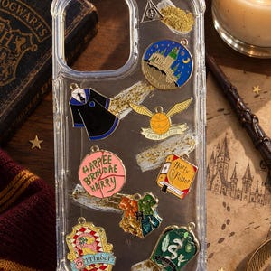 May include: A clear phone case decorated with various metal charms including a Hogwarts castle, a golden snitch with the text I OPEN AT THE CLOSE, a pink cake with HAPPEE B'RTHDAE HARRY, a Harry Potter book, house crests, and a deathly hallows symbol.