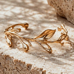 May include: A polished gold-tone cuff bracelet featuring an open design that mimics the organic shape of a slender branch. The metal structure includes a single prominent leaf element and several small, rounded bud-like protrusions along the band.