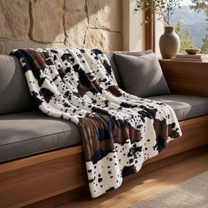 May include: A soft throw blanket featuring a white base with irregular brown and black cow-print spots. The blanket is draped over a gray sofa cushion, displaying a plush texture and a repeating pattern of dark patches across the entire surface.