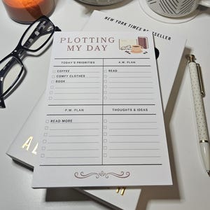 May include: A white daily planner notepad with the heading PLOTTING MY DAY, featuring sections for Today's Priorities, A.M. Plan, P.M. Plan, and Thoughts & Ideas. The notepad includes small illustrations of a book, glasses, and a coffee cup.