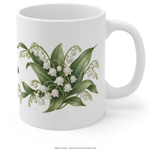 May include: A white ceramic mug featuring a botanical illustration of lily of the valley flowers with green leaves and small white bell-shaped blossoms. The design is centered on the front of the mug, which has a smooth finish and a curved handle.