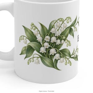 May include: A white ceramic mug featuring a detailed botanical illustration of lily of the valley flowers with green leaves and small white bell-shaped blossoms. The design is centered on the side of the mug, showcasing a cluster of stems.