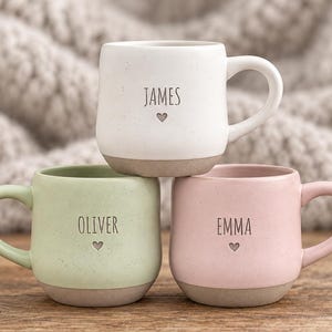 Puede incluir: Tres tazas de cerámica con acabados moteados en blanco, verde claro y rosa claro. Cada taza presenta un nombre impreso en negro sobre un pequeño icono de corazón. Los nombres son James en la taza blanca, Oliver en la verde y Emma en la rosa.