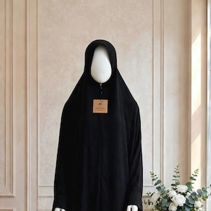 Può includere: Un'abaya nera lunga fino ai piedi con maniche lunghe e copricapo integrato. L'indumento è esposto su un manichino e presenta una piccola etichetta rettangolare marrone attaccata sul petto con del testo visibile.