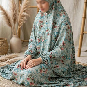May include: A light blue prayer garment featuring a floral pattern with pink and dark blue flowers. The one-piece outfit includes an integrated head covering, long sleeves with elastic cuffs, and a front placket with three white buttons.