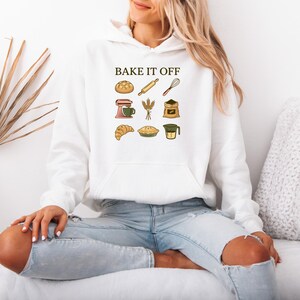 May include: A white hooded sweatshirt featuring the text BAKE IT OFF in dark brown letters above a grid of nine colorful baking-themed illustrations, including a loaf of bread, rolling pin, whisk, stand mixer, wheat, flour, croissant, pie, and measuring cup.