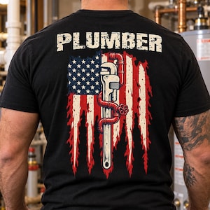 May include: A black t-shirt featuring the word PLUMBER in white block letters above a graphic of a distressed American flag. A silver pipe wrench is positioned vertically over the flag, with red plumbing pipes wrapped around the wrench handle.