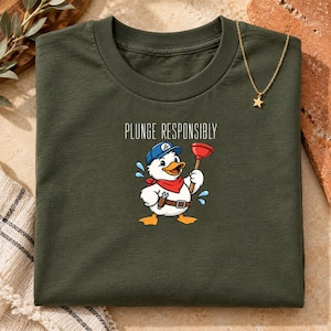 May include: A dark olive green t-shirt featuring a graphic of a white duck wearing a blue cap and red neckerchief, holding a red plunger. The text "PLUNGE RESPONSIBLY" is printed above the duck, which also wears a brown tool belt.