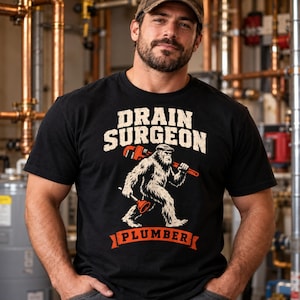 May include: A black t-shirt featuring a graphic of a hairy creature wearing a cap, carrying a large red pipe wrench on its shoulder and holding a red plunger. The text DRAIN SURGEON is printed above the creature, and PLUMBER is below.