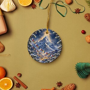 May include: A round ceramic ornament featuring a snowy mountain landscape at night with a bright star above the peak and colorful prayer flags strung across the scene. The ornament is suspended by a thin gold cord against a solid tan background.