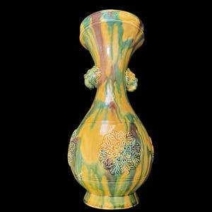 Może przedstawiać: Ceramiczny wazon z wykończeniem w żółte, zielone i brązowe zacieki szkliwa. Wazon posiada rozszerzany wylot, wąską szyjkę z dwoma małymi uchwytami w kształcie lwich głów po bokach oraz pękaty korpus ozdobiony tłoczonymi kwiatowymi medalionami.