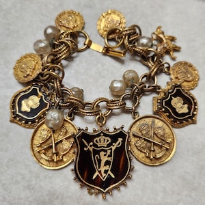 Puede incluir: Una pulsera de dijes en tono dorado con múltiples colgantes, incluyendo monedas redondas, dijes en forma de escudo con esmalte negro y perlas de imitación color crema. El escudo central muestra un emblema dorado de león, corona y espada.