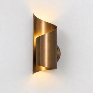 Puede incluir: Un aplique de pared con una pantalla cilíndrica y curva de metal latón con acabado cepillado. El diseño consta de dos paneles verticales superpuestos que emiten un brillo cálido y suave a través de las aberturas superior e inferior.