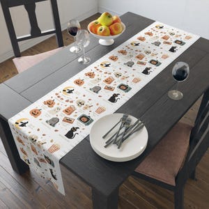 Puede incluir: Un camino de mesa blanco con un patrón repetitivo de ilustraciones temáticas de Halloween que incluyen calabazas, gatos negros, fantasmas, lápidas, calaveras, palomitas, casetes y televisores antiguos, cada uno con un pequeño texto en negro.
