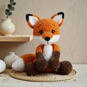 Puede incluir: Un peluche de zorro tejido a ganchillo con hilo naranja, blanco y marrón oscuro. El zorro tiene grandes ojos negros, una pequeña nariz negra, orejas puntiagudas con centros blancos y una cola tupida con punta blanca, sentado.