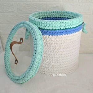 Puede incluir: Una cesta cilíndrica de ganchillo con base blanca, una banda central azul y un borde verde menta. La tapa a juego presenta un borde de ganchillo verde menta, un centro transparente y un asa de cuero marrón. El texto @artesbabylu es visible.