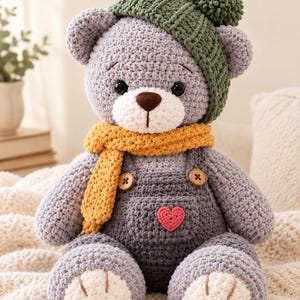 Puede incluir: Un oso de peluche tejido a ganchillo de color gris que lleva un gorro de punto verde con pompón, una bufanda amarilla y un peto gris oscuro. El peto tiene dos botones de madera y un pequeño parche de corazón rosa.