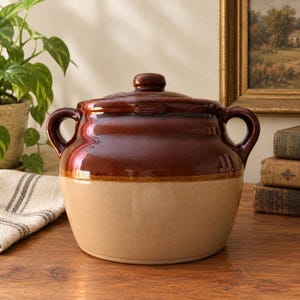 May include: A two-handled ceramic crock with a lid, featuring a glossy dark brown glaze on the upper portion and a smooth, matte beige finish on the lower half. The vessel has a rounded body, a small knob on the lid, and two side handles.